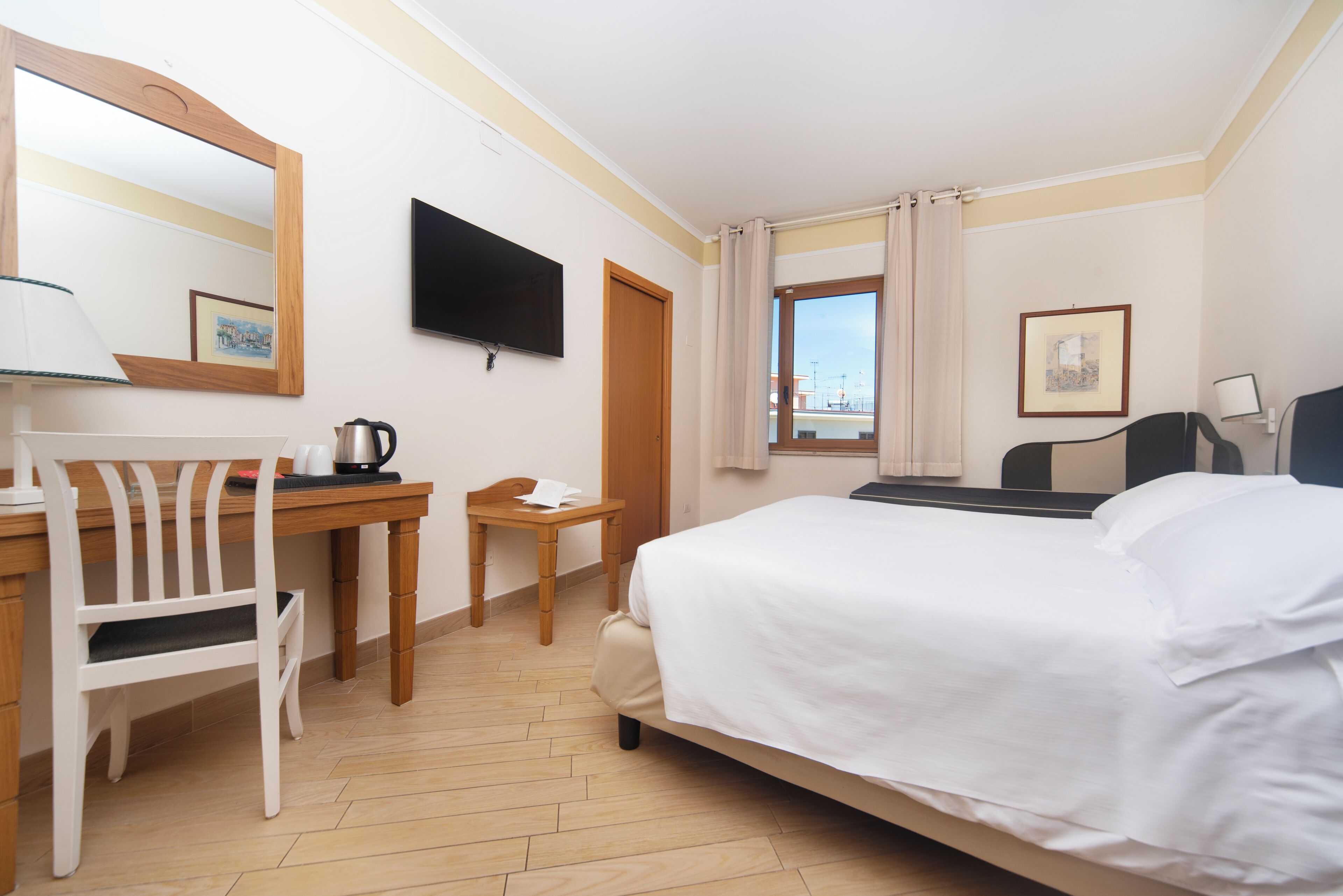 A hotel room with a bed, a desk, a chair, a TV, a window with curtains, and a framed picture on the wall.