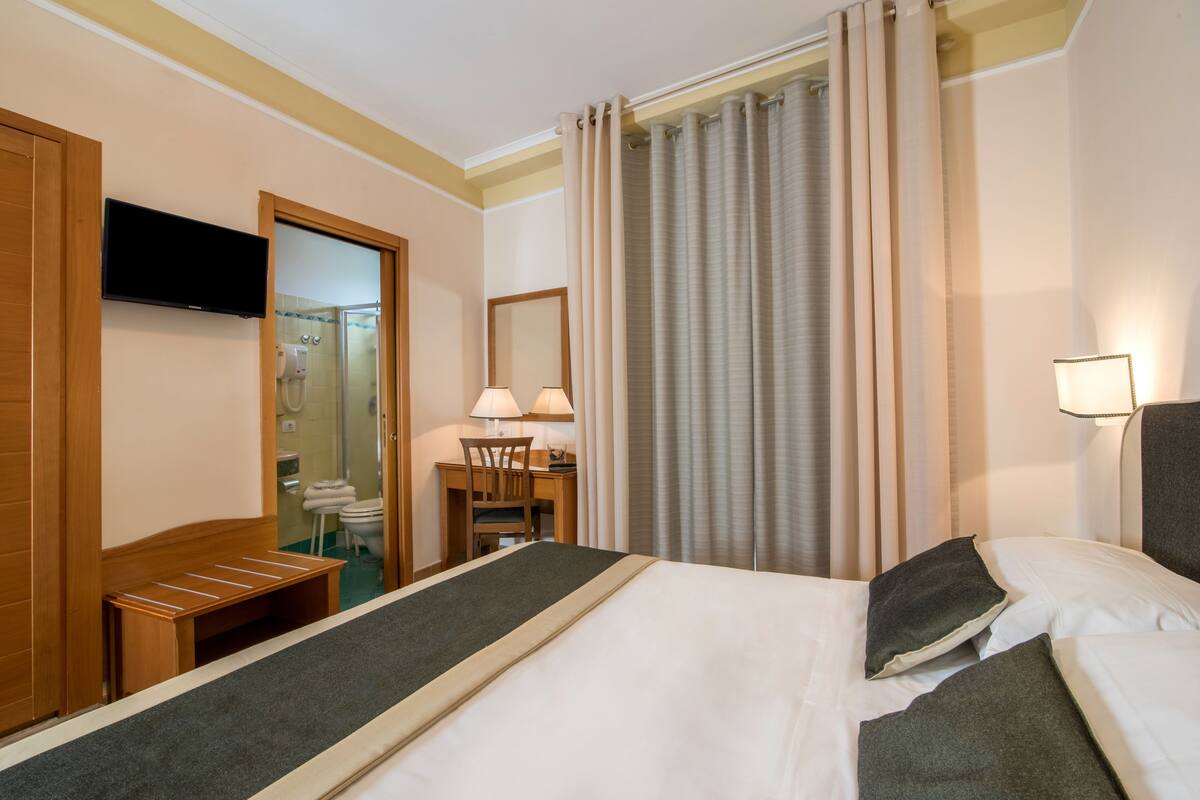 A hotel room with a bed, a desk with a chair, a television, a bathroom with a shower, and a window with curtains.