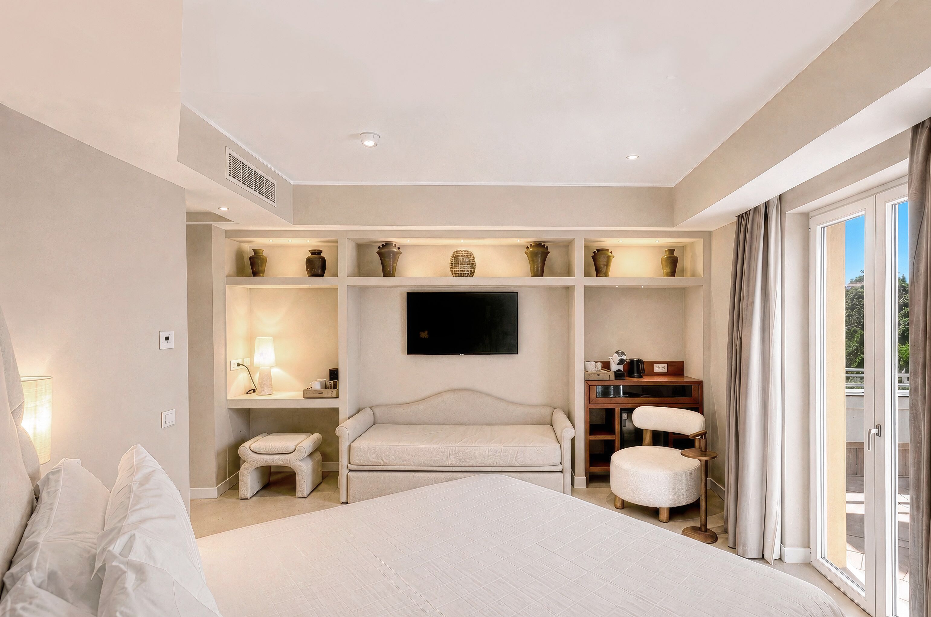 A hotel room with a large bed, a flat-screen TV, a seating area with a sofa and chairs, and a balcony with curtains.
