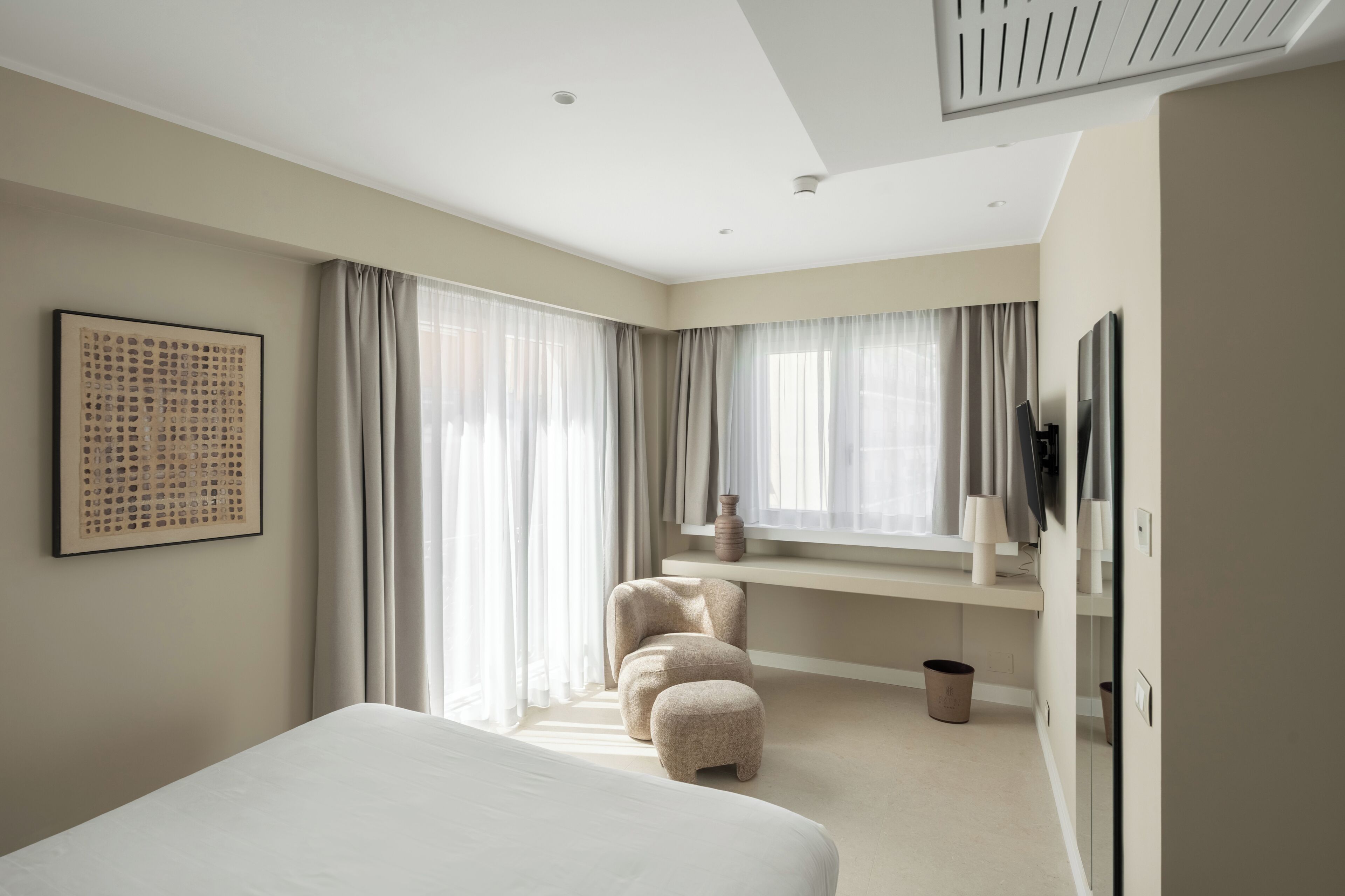 A modern hotel room with a large bed, a flat-screen TV, a seating area with ottomans, and a large window with sheer curtains.