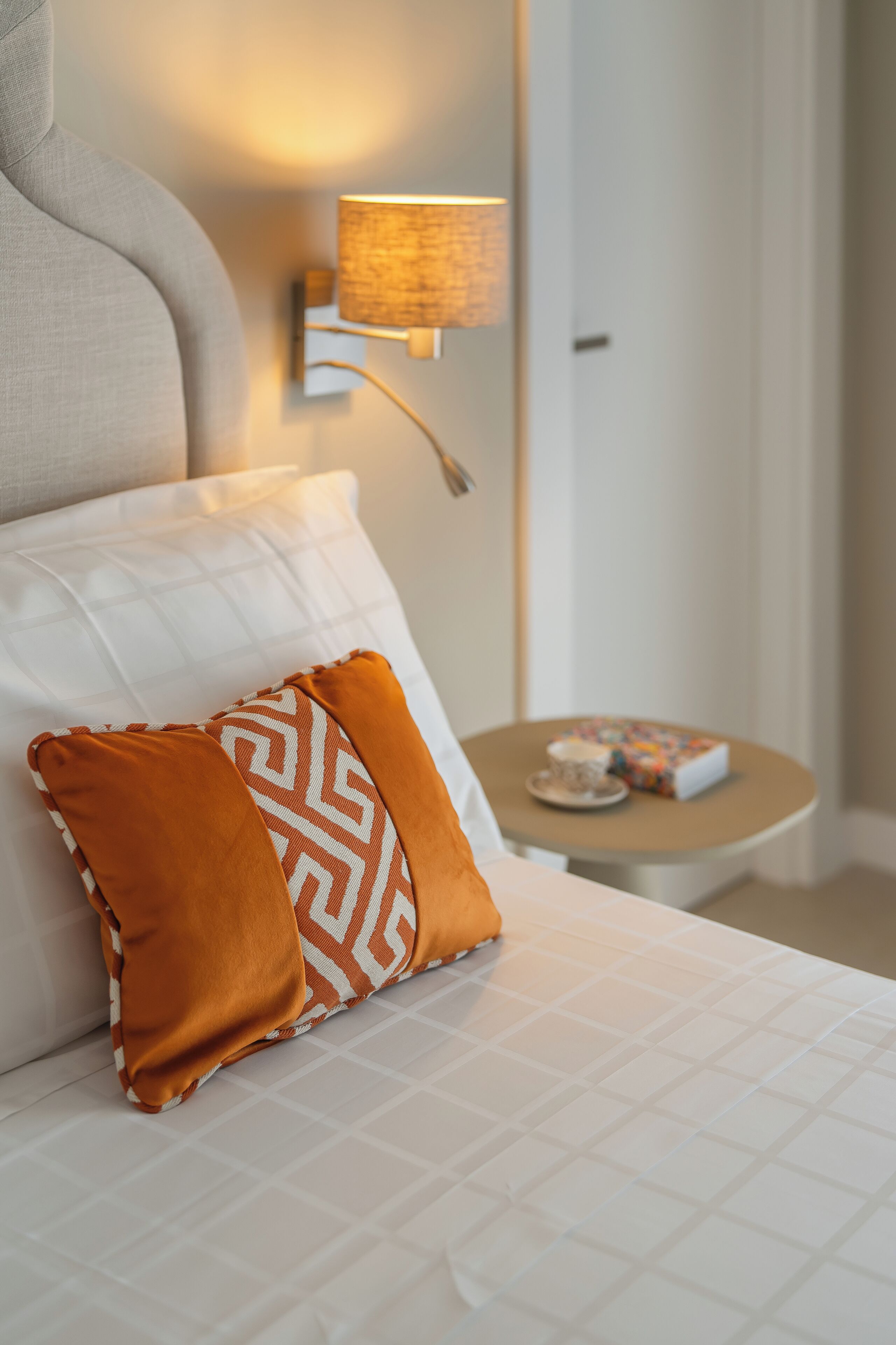 Een keurig opgemaakt bed met witte lakens en twee decoratieve kussens, één oranje en één wit met een patroon. Een wandlamp met een gestructureerde kap is zichtbaar, en in de achtergrond staat een klein rond tafeltje met een kop en een doos.
