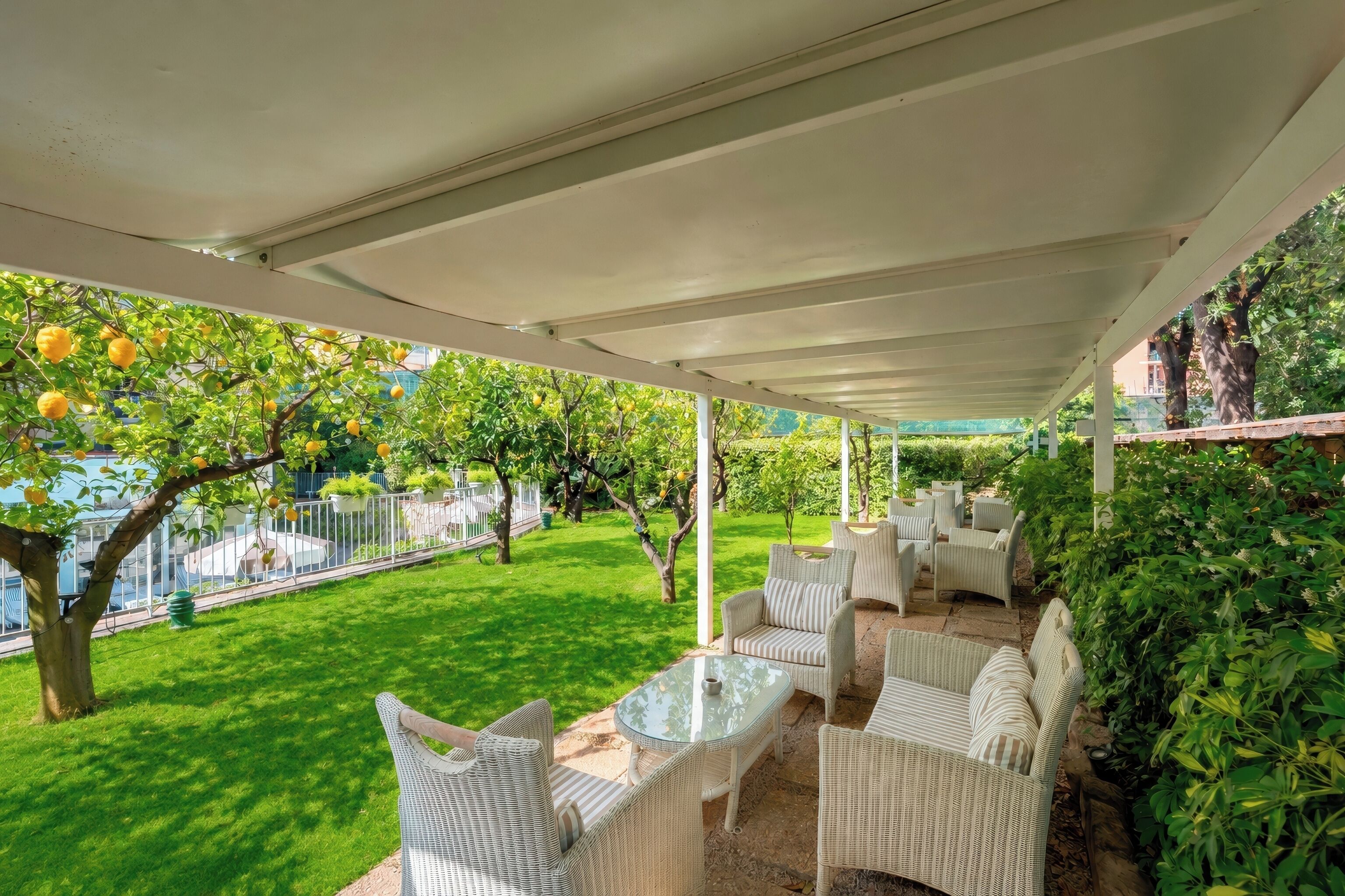 Una veranda esterna con mobili in vimini e un tavolo in vetro, circondata da un rigoglioso giardino e alberi da frutto.
