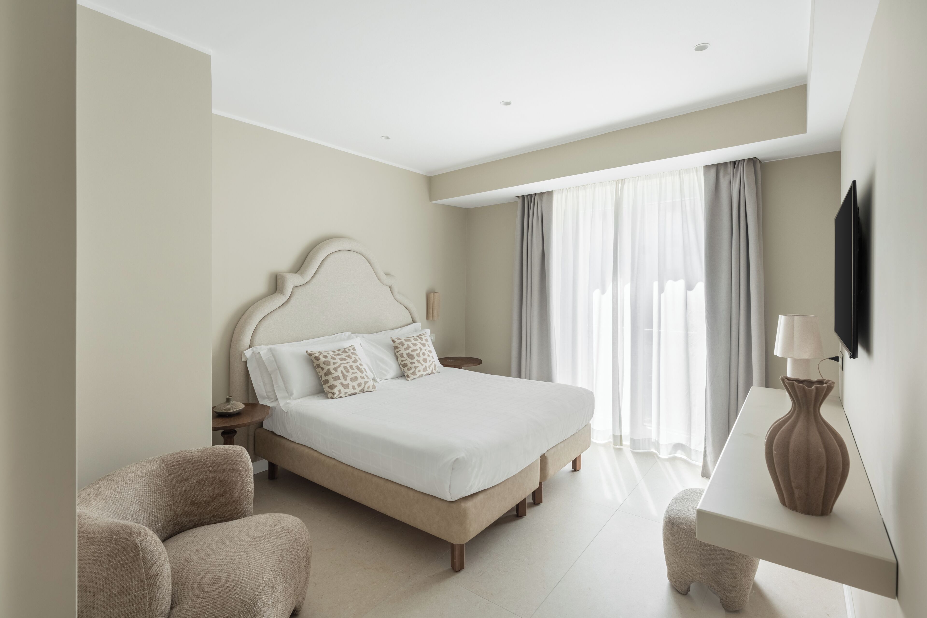 A hotel room with a bed, a chair, a bedside table, a lamp, and a TV mounted on the wall.