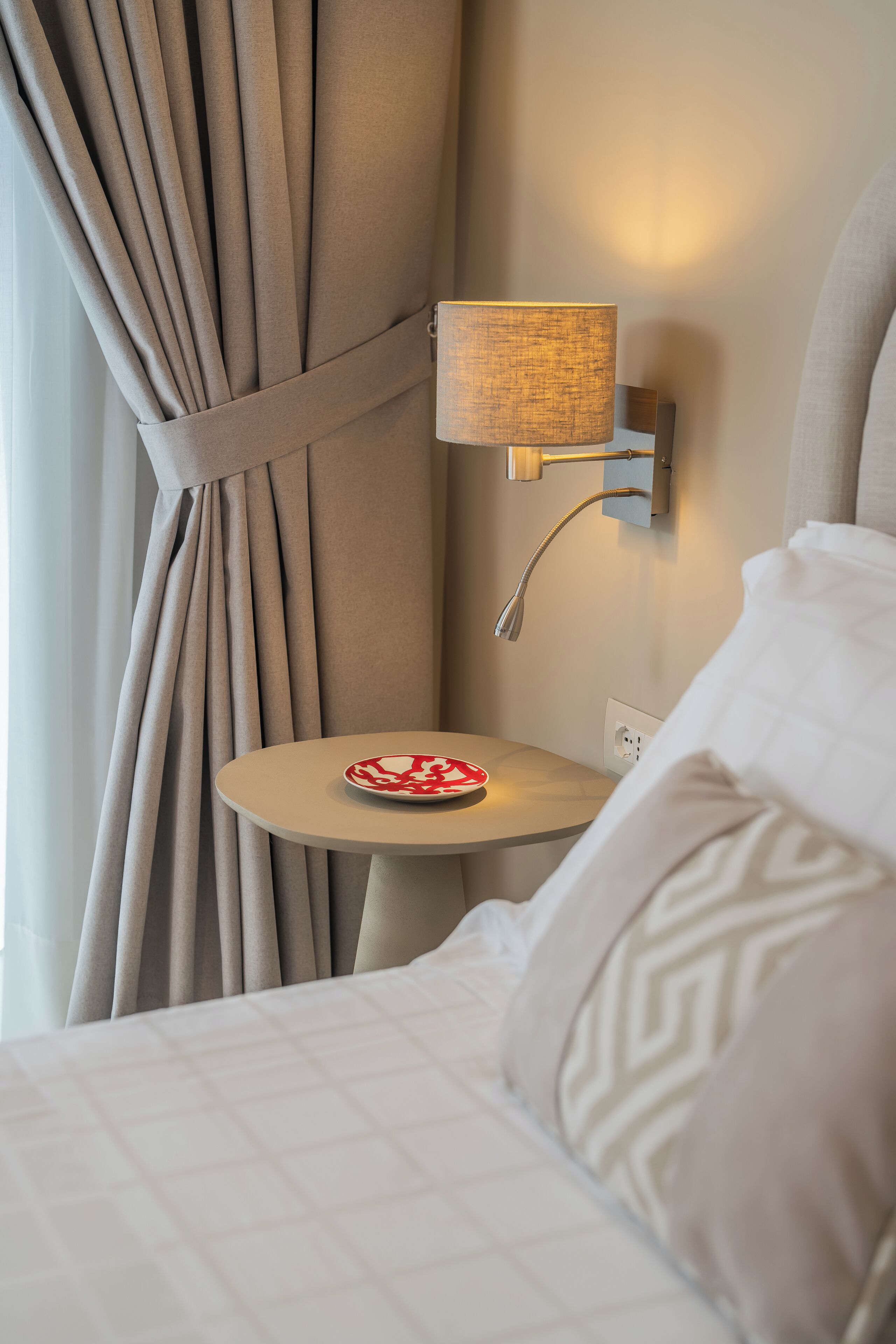 A hotel room with a bed, a bedside table with a lamp, and a view through curtains.
