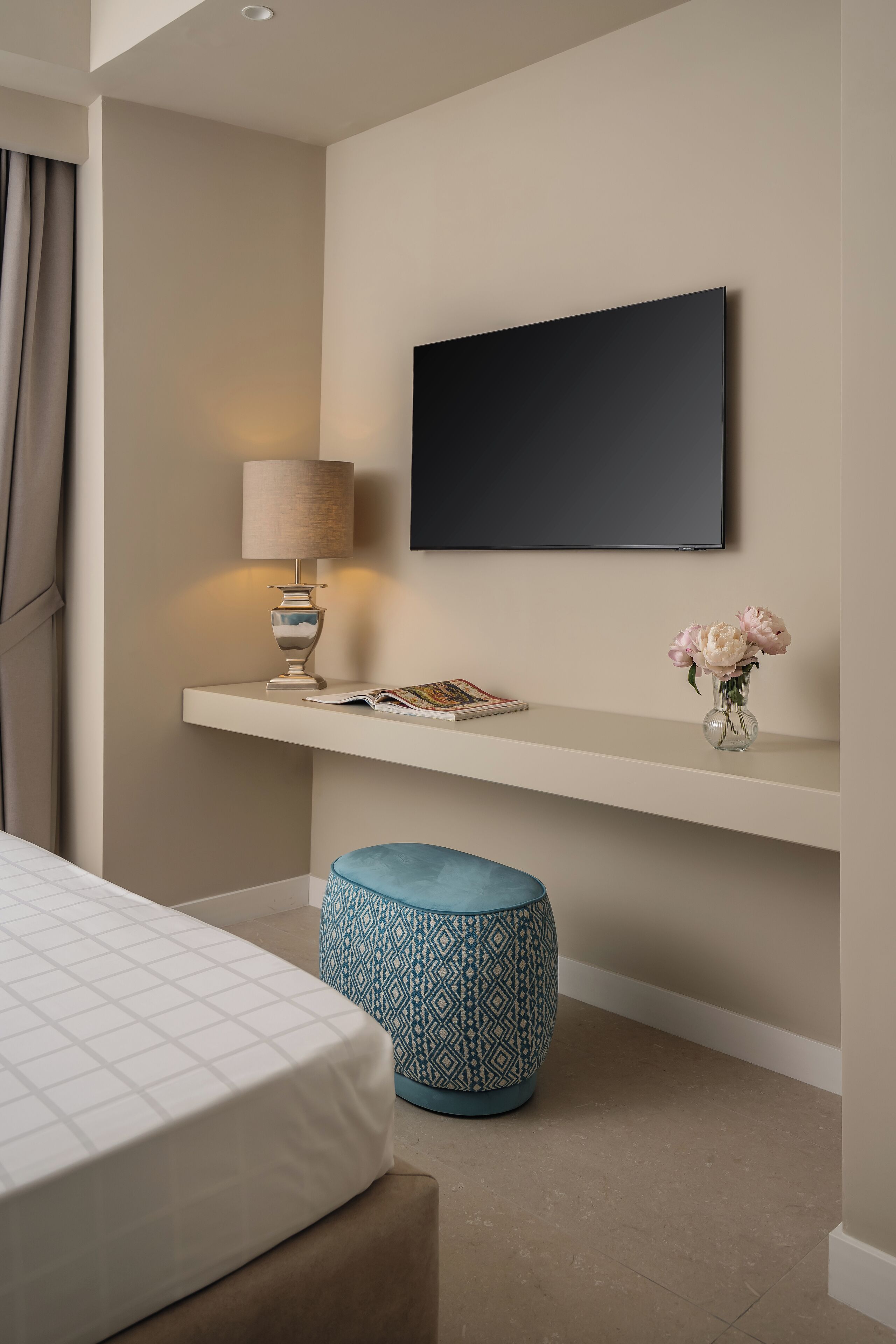 A modern hotel room with a bed, a bedside table with a lamp, a TV mounted on the wall, a small shelf with a vase of flowers, and a patterned ottoman.