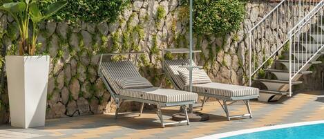 A poolside area with lounge chairs and a covered umbrella, a stone wall with greenery, and a staircase.