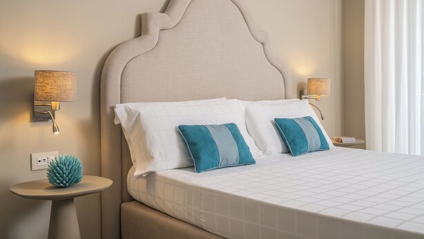 A neatly made bed with a headboard, two bedside lamps, and a small table with a decorative item. - Hotel Capri - Adult Only (Sorrento)