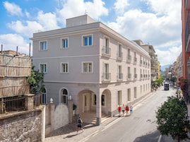 Un hotel a più piani con un ingresso ben visibile, un vialetto recintato e una vista della strada con persone che passeggiano.