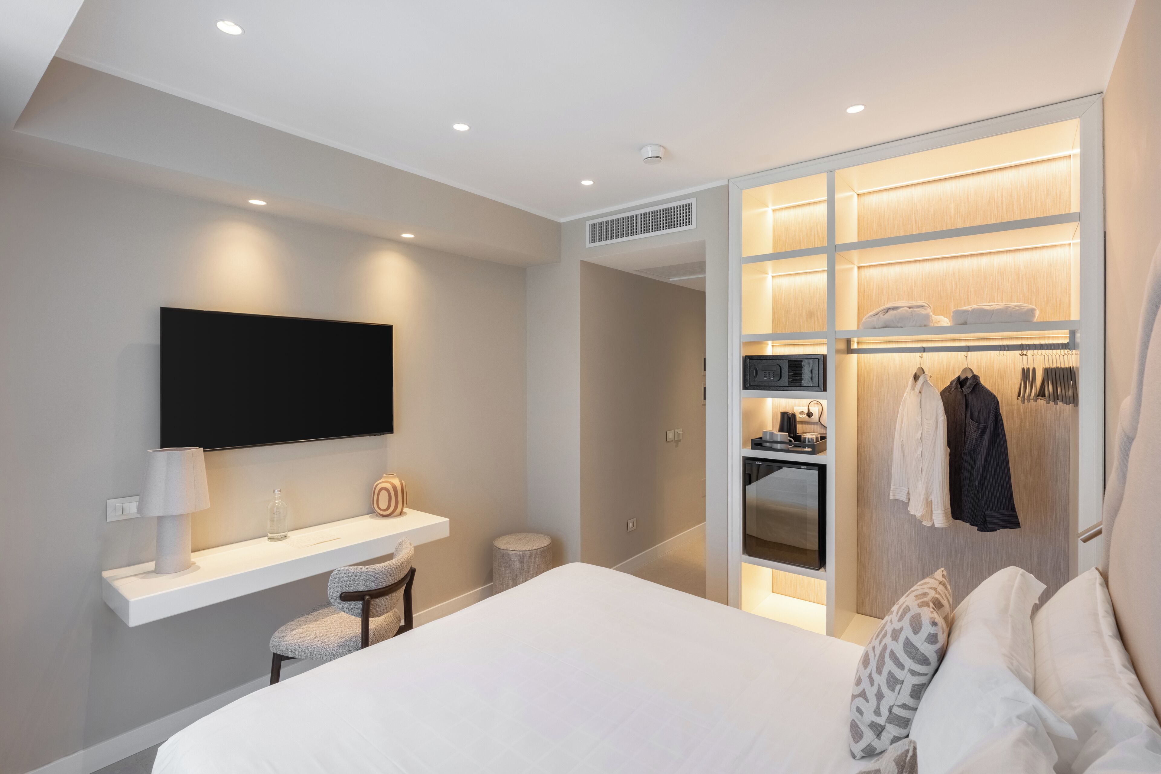 A modern hotel room with a bed, a TV mounted on the wall, a vanity with a mirror, and a walk-in closet with clothing and a microwave.