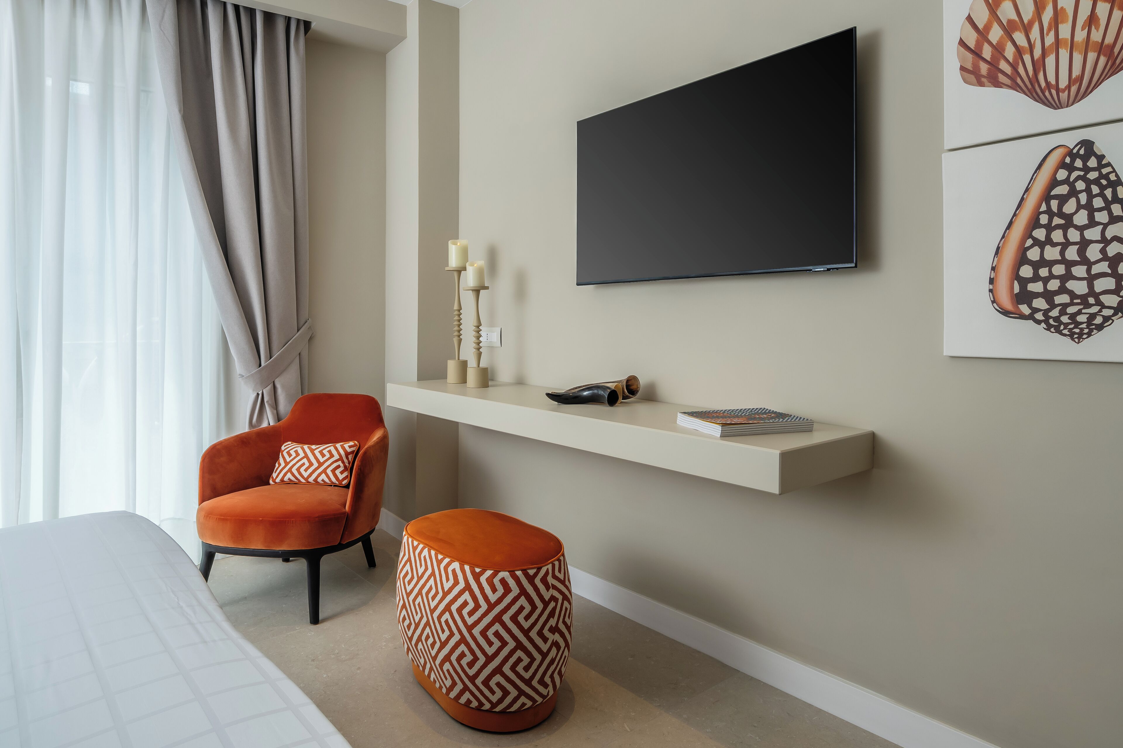 A modern hotel room with a bed, a TV mounted on the wall, a small seating area with a chair and ottoman, and a shelf with decorative items.