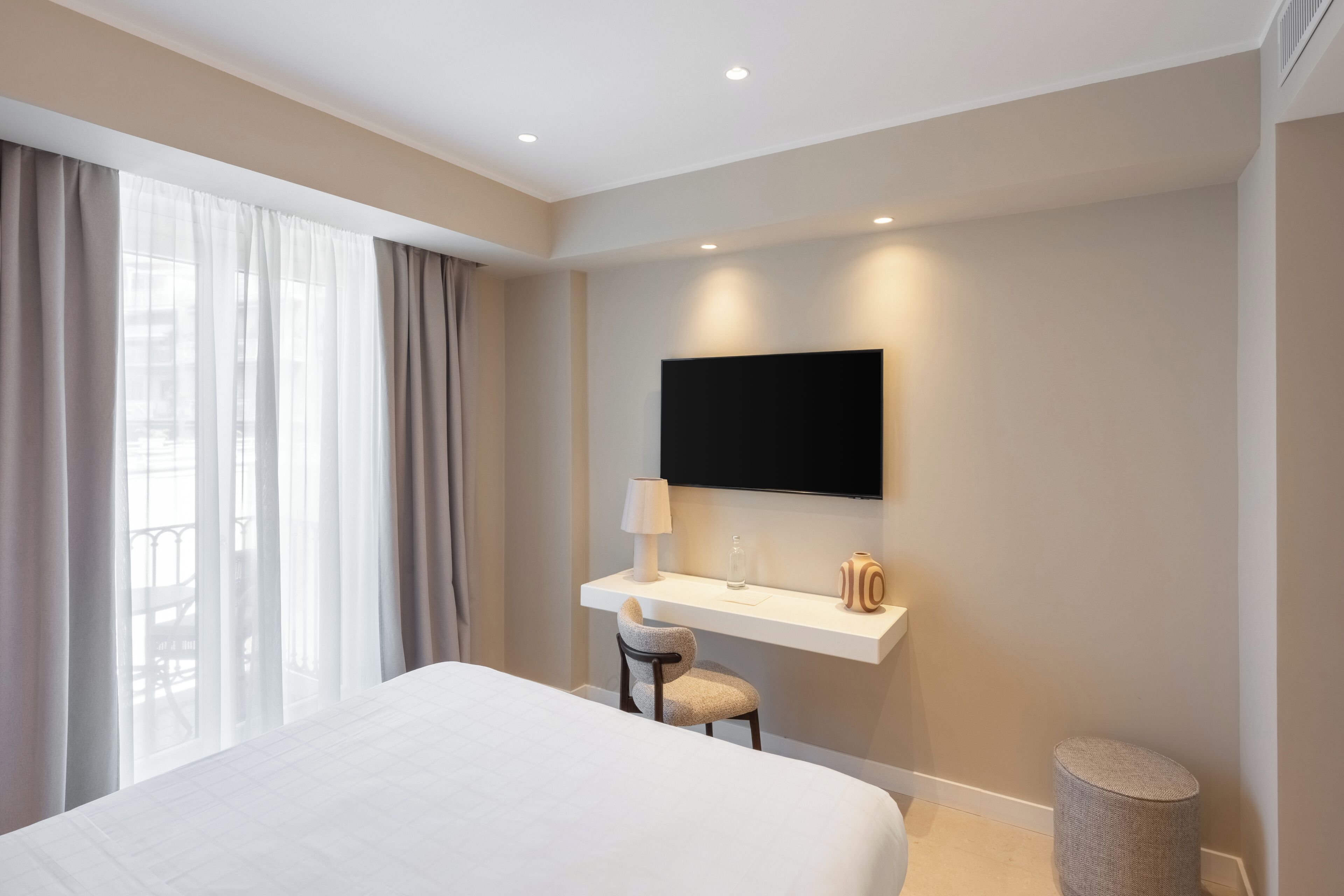 Une chambre d’hôtel moderne dotée d’un grand lit, d’un téléviseur fixé au mur, d’un petit bureau avec une lampe et offrant une vue sur l’extérieur à travers des rideaux légers.