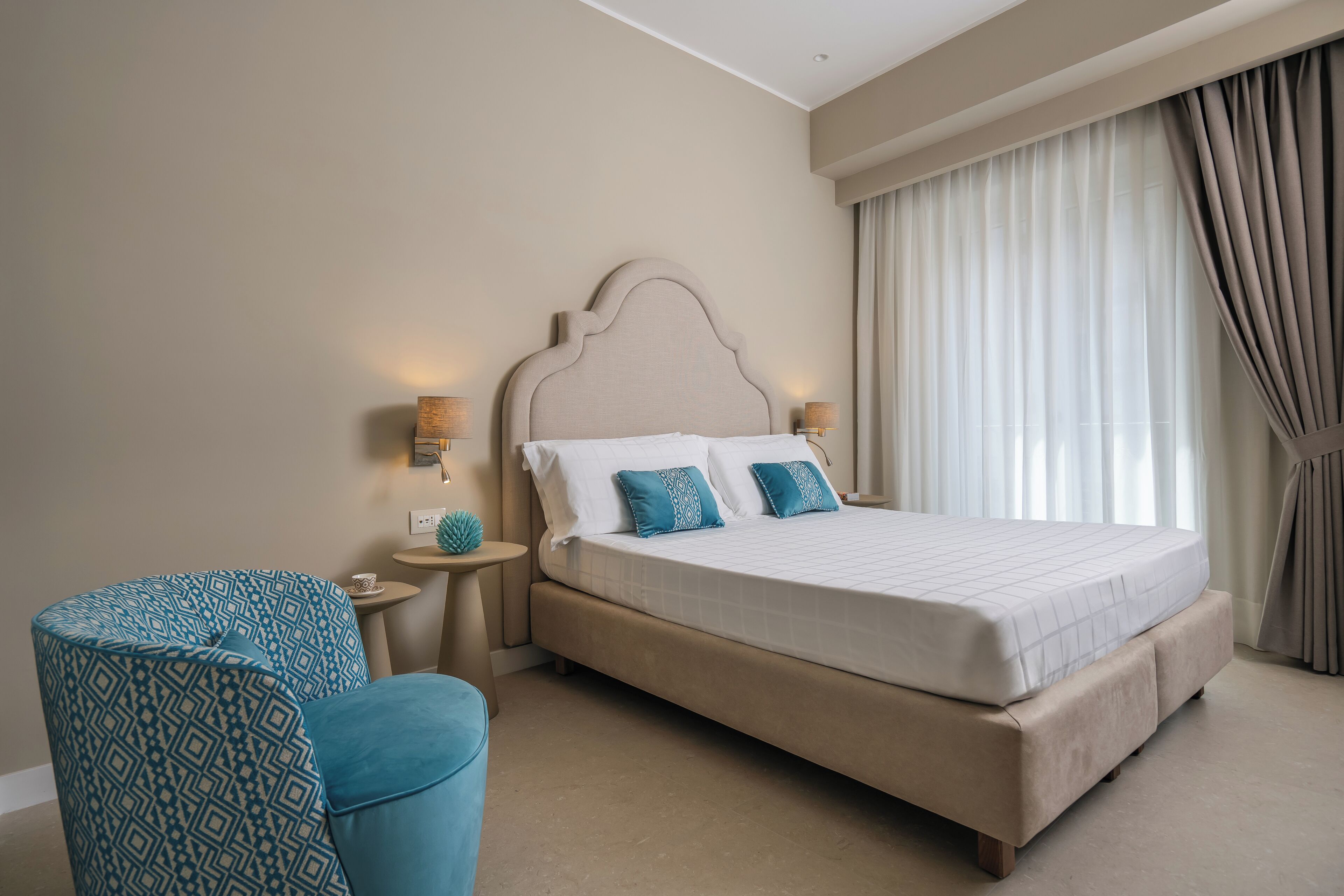 A bedroom with a large bed, a round blue chair, two bedside tables with lamps, and a window with curtains.