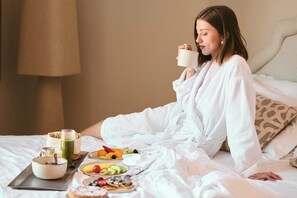 Une personne vêtue d’un peignoir blanc est assise sur un lit, et devant elle se trouvent des aliments pour le petit-déjeuner, notamment des fruits, des crêpes et une tasse.