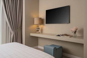 Habitación de hotel con una cama, una mesita de noche con lámpara, un televisor de pantalla plana, una estantería con un libro y un jarrón de flores.