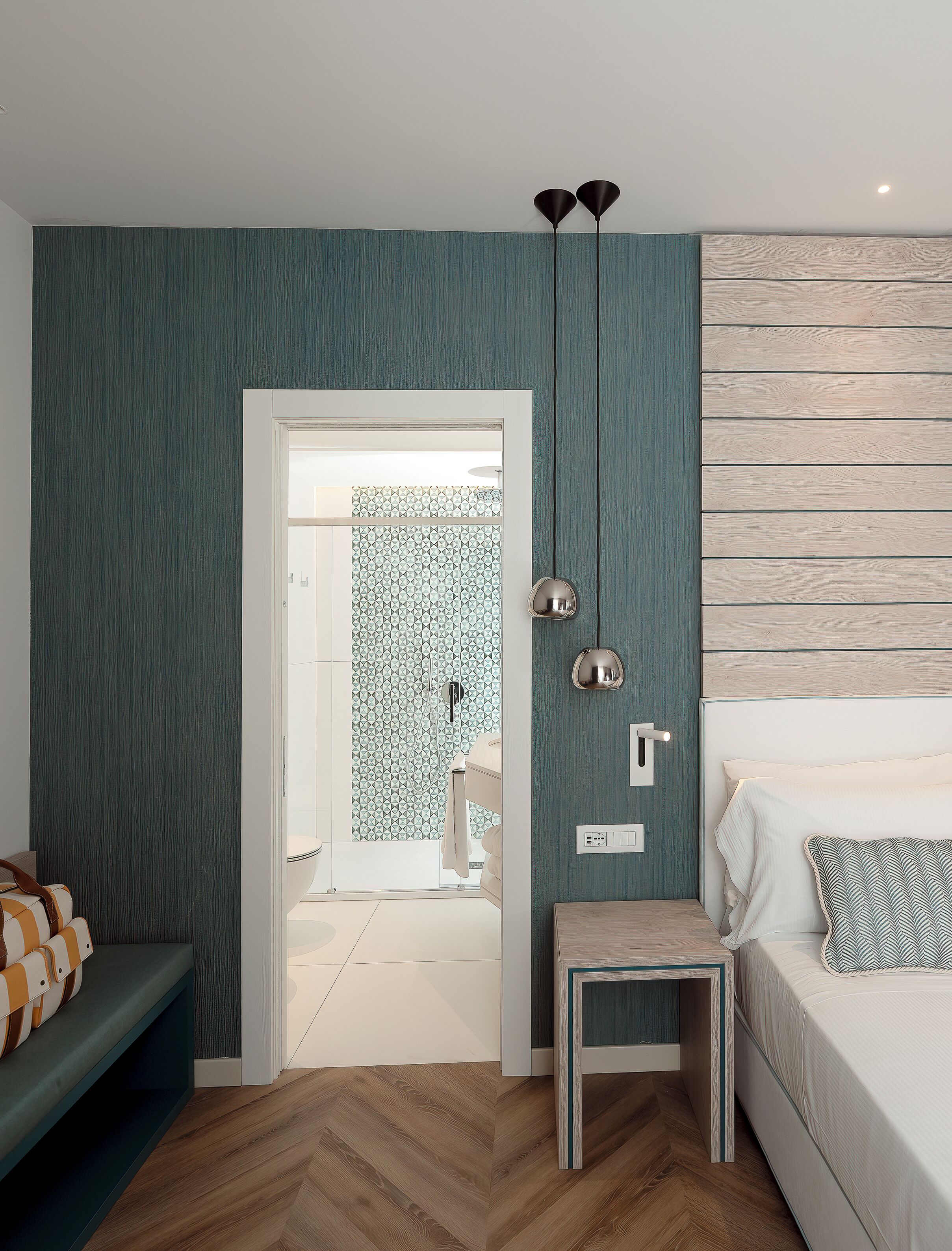 Une chambre moderne avec un parquet en bois à motifs en chevrons, un lit agrémenté d’un coussin à motifs, une table de chevet et une lampe fixée au mur.