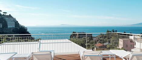 Una terraza con sillas y mesas de metal blanco, con vistas a la costa, edificios y el mar.