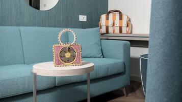 Ein salbeigrünliches Sofa mit einem runden Couchtisch davor. Drei Wandspiegel und eine Korbtasche auf einem kleinen Tisch sind zu sehen.