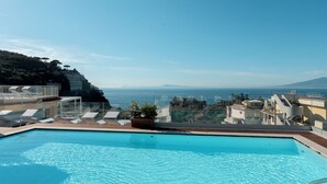 Piscina azul cristalina com espreguiçadeiras e um vaso de planta, com vista para a costa, edifícios e uma cordilheira ao longe.