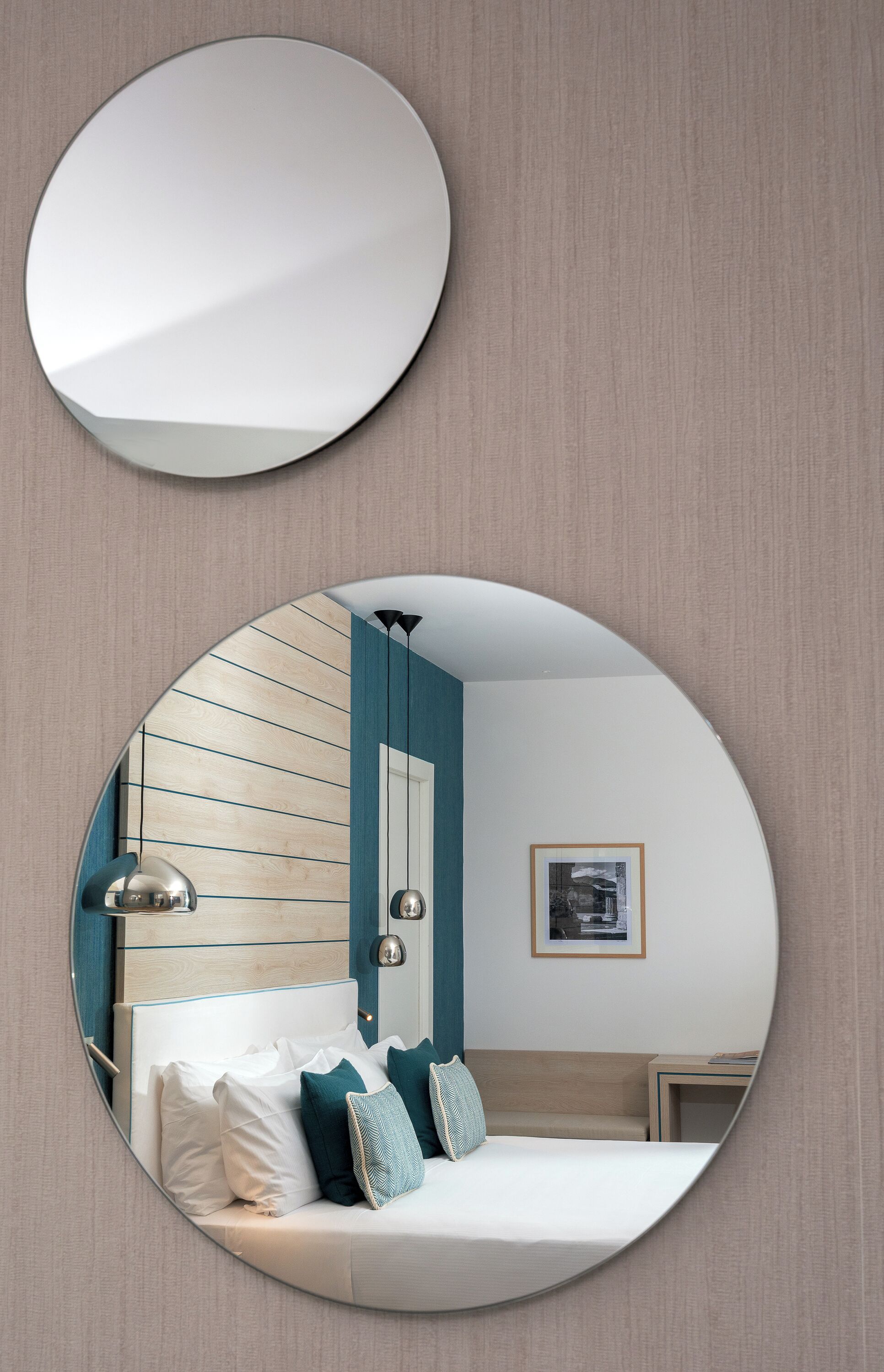 Un espejo circular refleja un dormitorio con cama, almohadas, una mesita de noche y un cuadro en la pared.