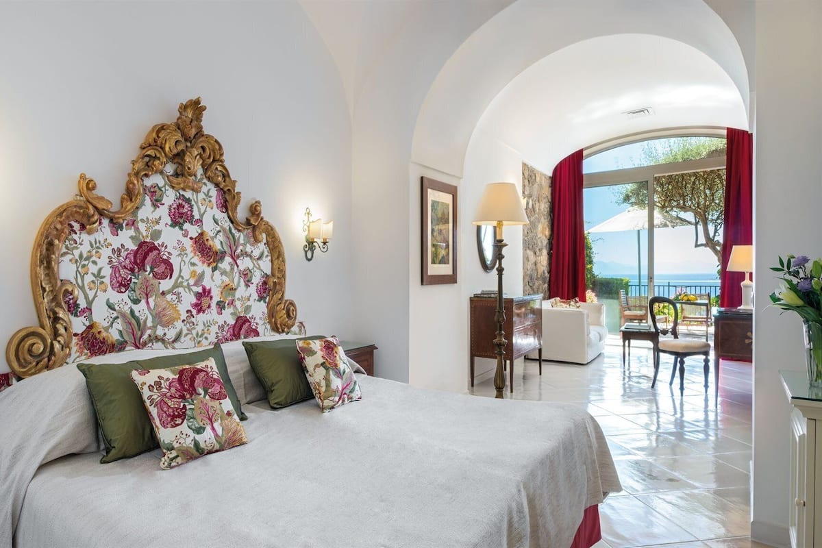 Ein Hotelzimmer mit einem großen Bett, einem blumengemusterten Kopfteil, zwei Wandleuchten, Blick ins Freie und einem Balkon mit Tisch und Stühlen.