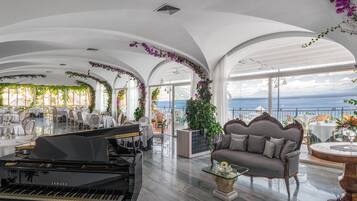 A spacious, elegantly decorated lounge with a grand piano, comfortable seating, and a view of the sea.