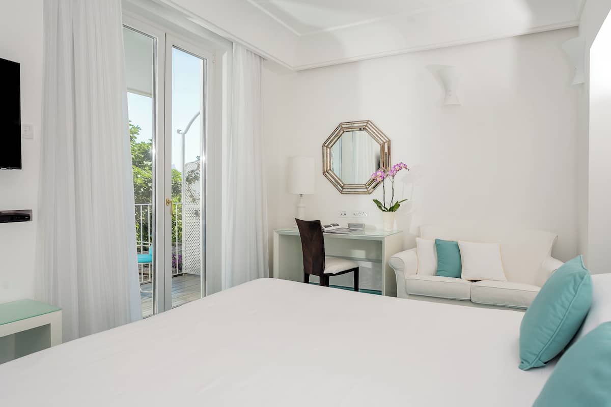 Ein Hotelzimmer mit Bett, Schreibtisch, Sessel, Sofa, Fernseher und einem Balkon mit Vorhängen.
