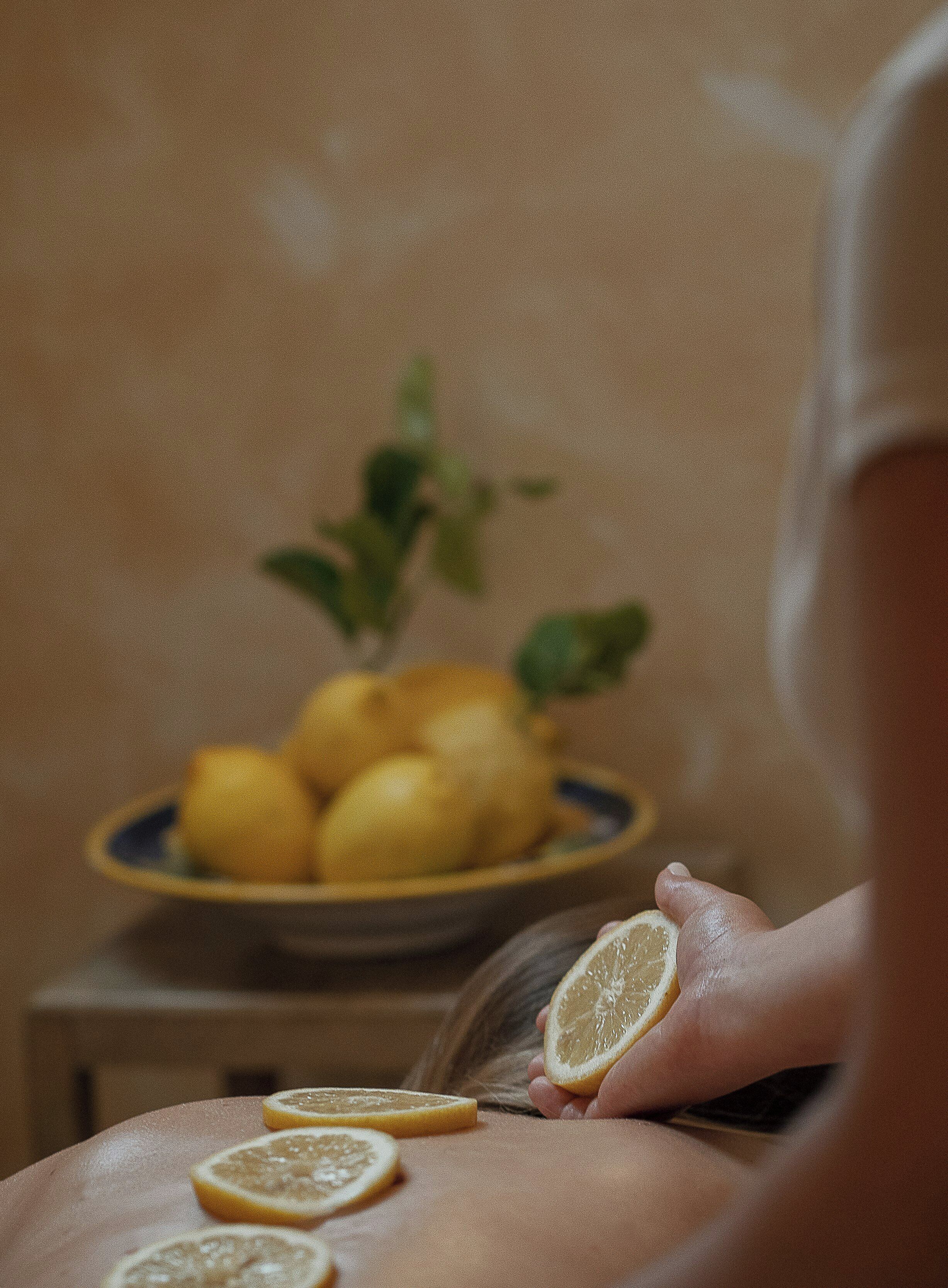 A close-up of a hand holding a lemon slice with a plate of lemons in the background.