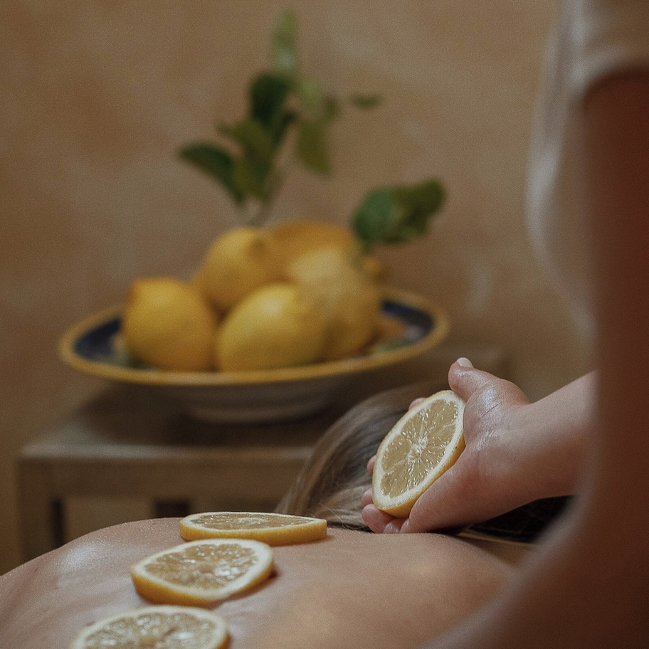 A close-up of a hand holding a lemon slice with a plate of lemons in the background.