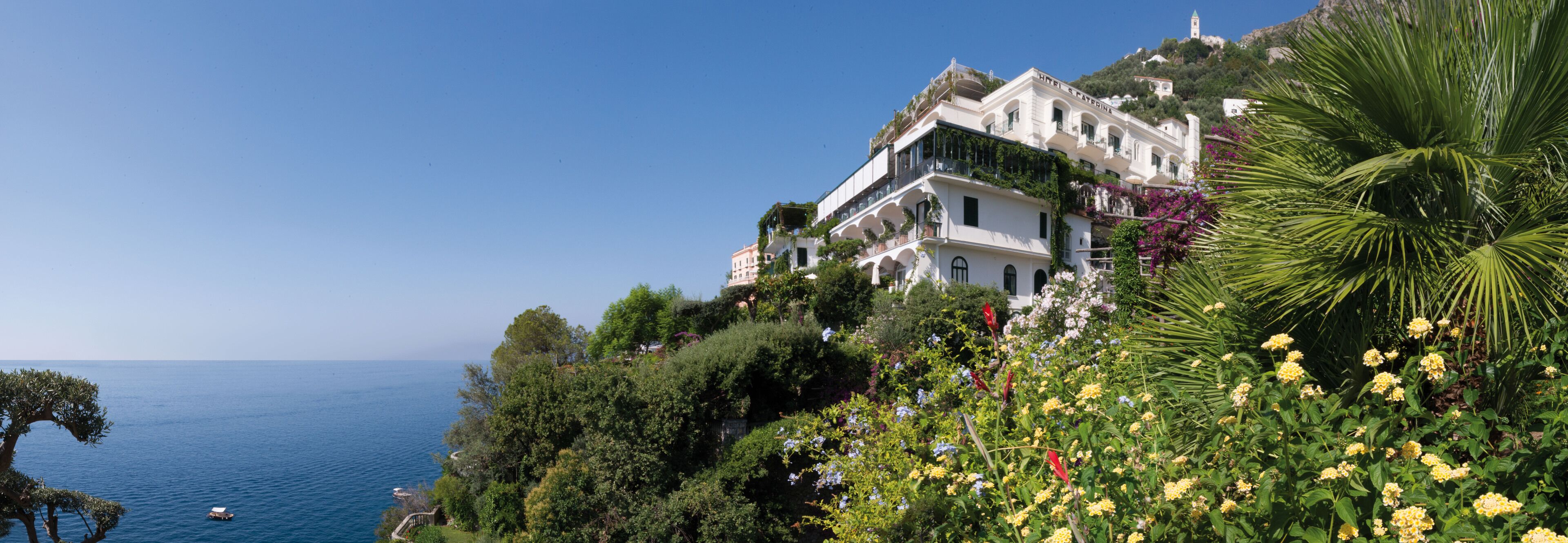 Ein Küstenhotel mit mehreren Balkonen mit Blick auf das Meer, umgeben von üppiger Vegetation und blühenden Pflanzen.
