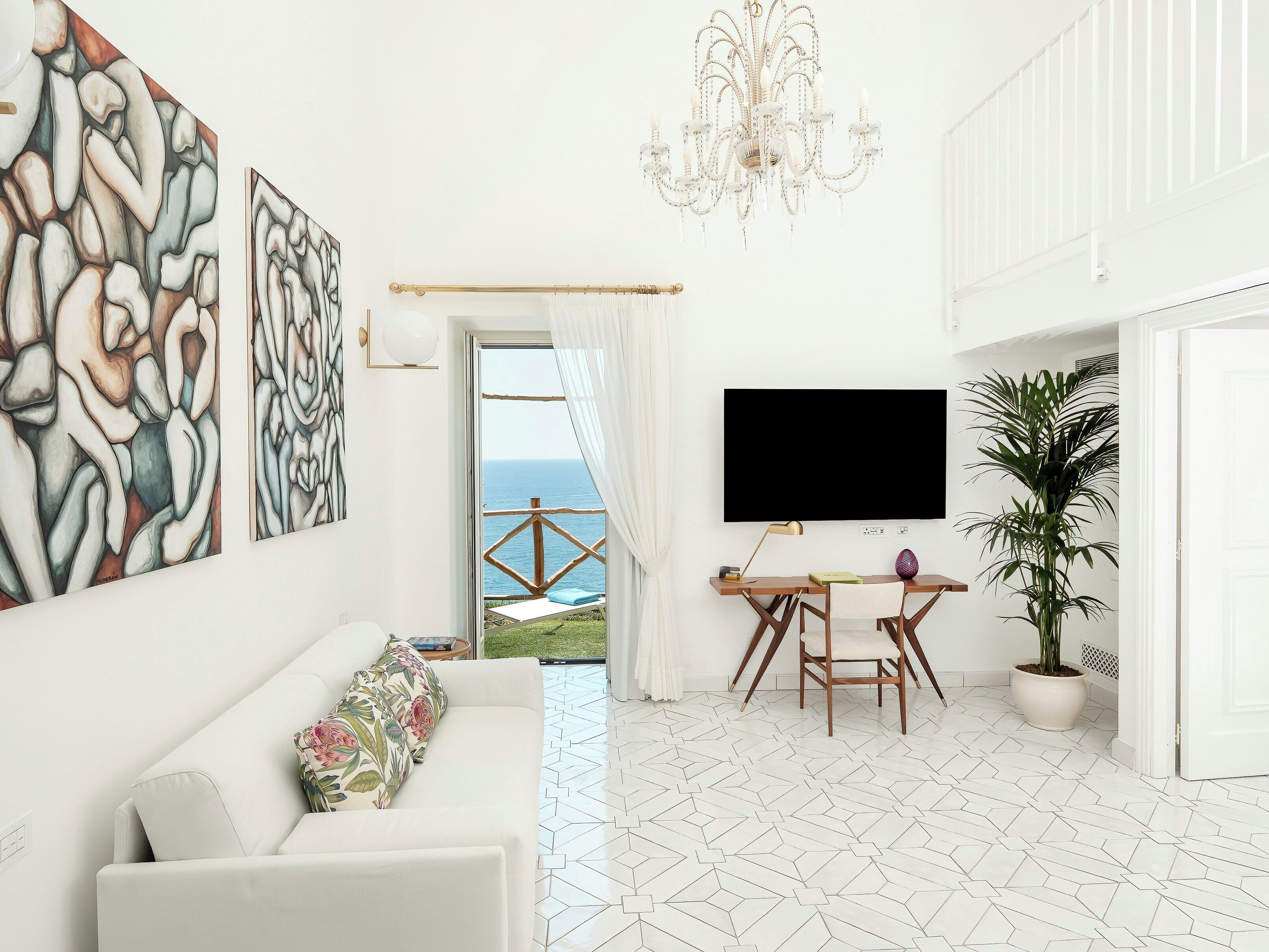 A modern living room with a white sofa, abstract wall art, a chandelier, a flat-screen TV, a dining table with chairs, and a potted plant.