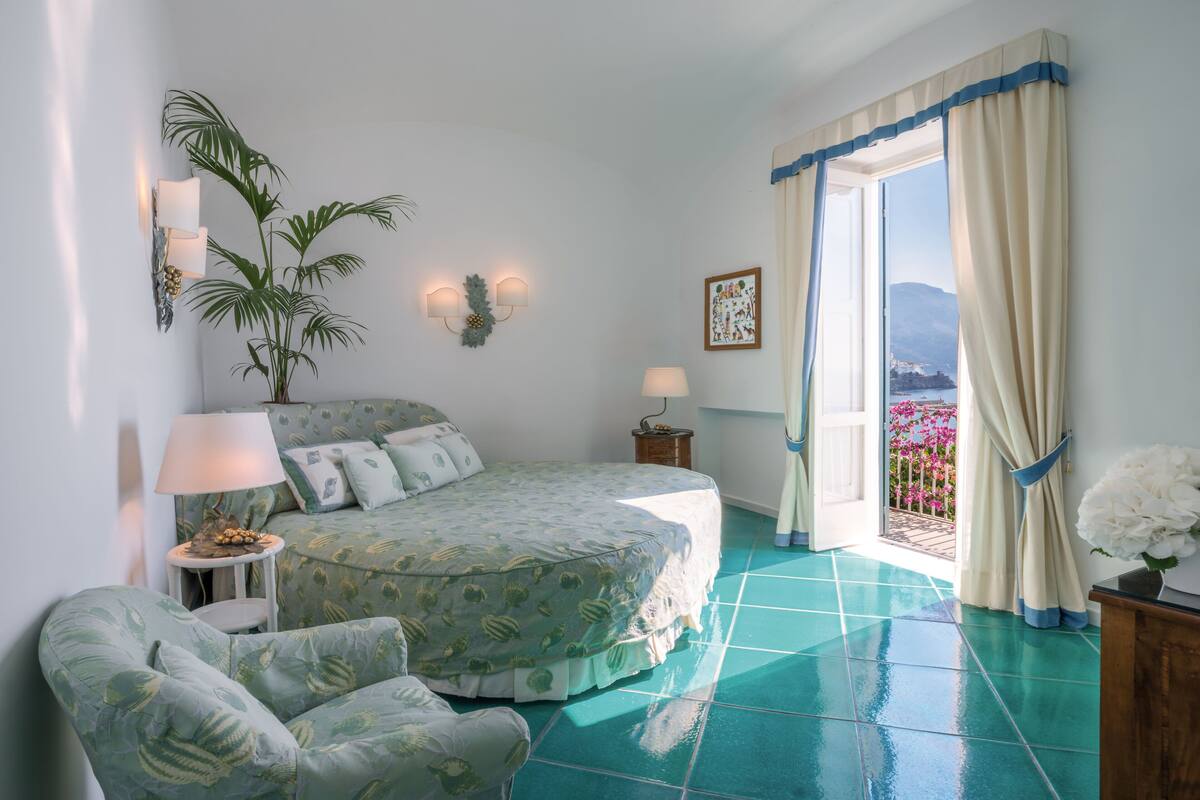 Ein Schlafzimmer mit Bett, Stuhl, Nachttisch, Lampe, Zimmerpflanze und Blick ins Freie.