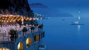 Un restaurante frente al mar con terraza, un edificio de piedra con arcos y barcos al fondo.