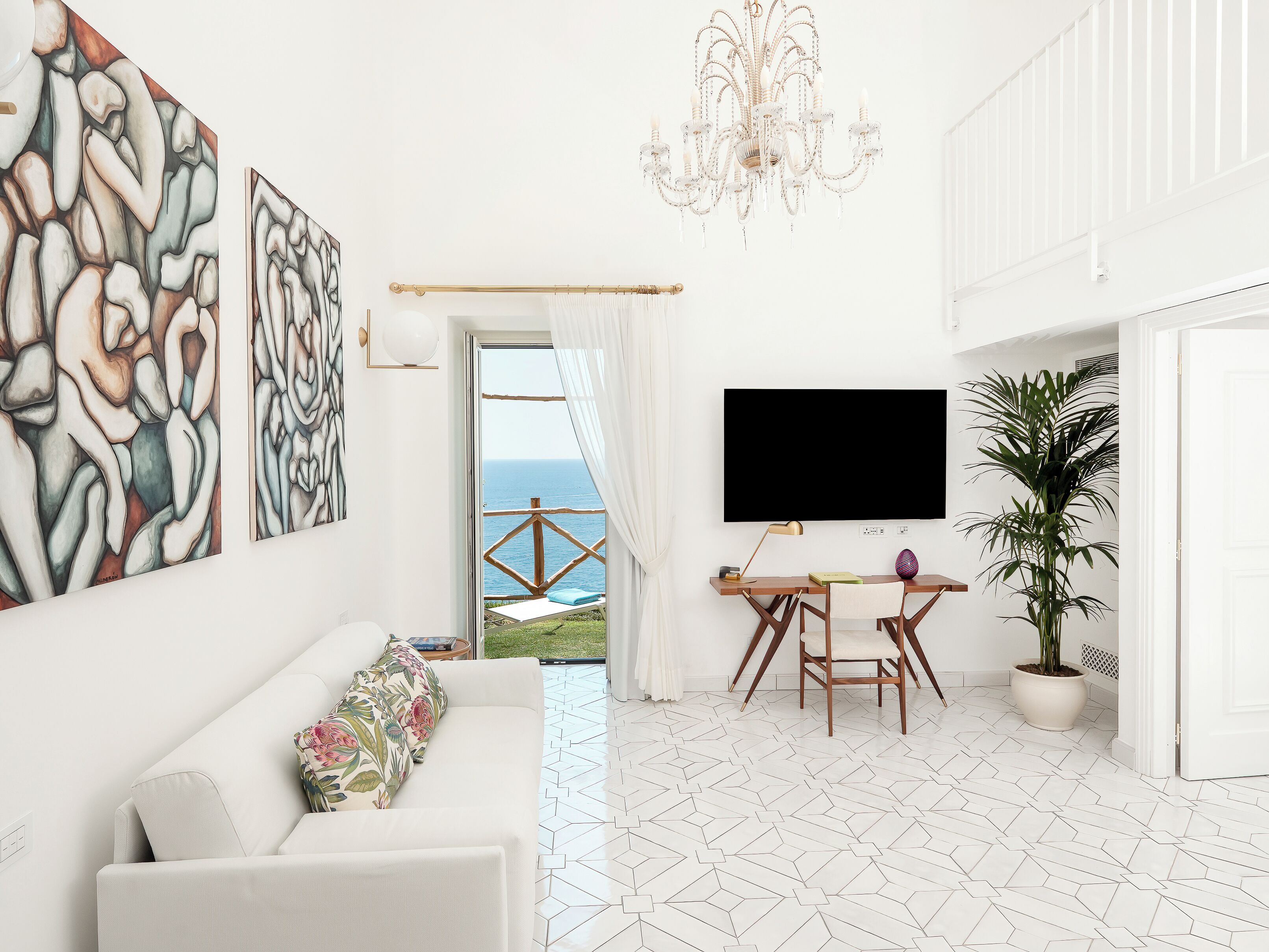 A modern living room with a white sofa, abstract wall art, a chandelier, a flat-screen TV, a dining area with a wooden table and chairs, and a potted plant.