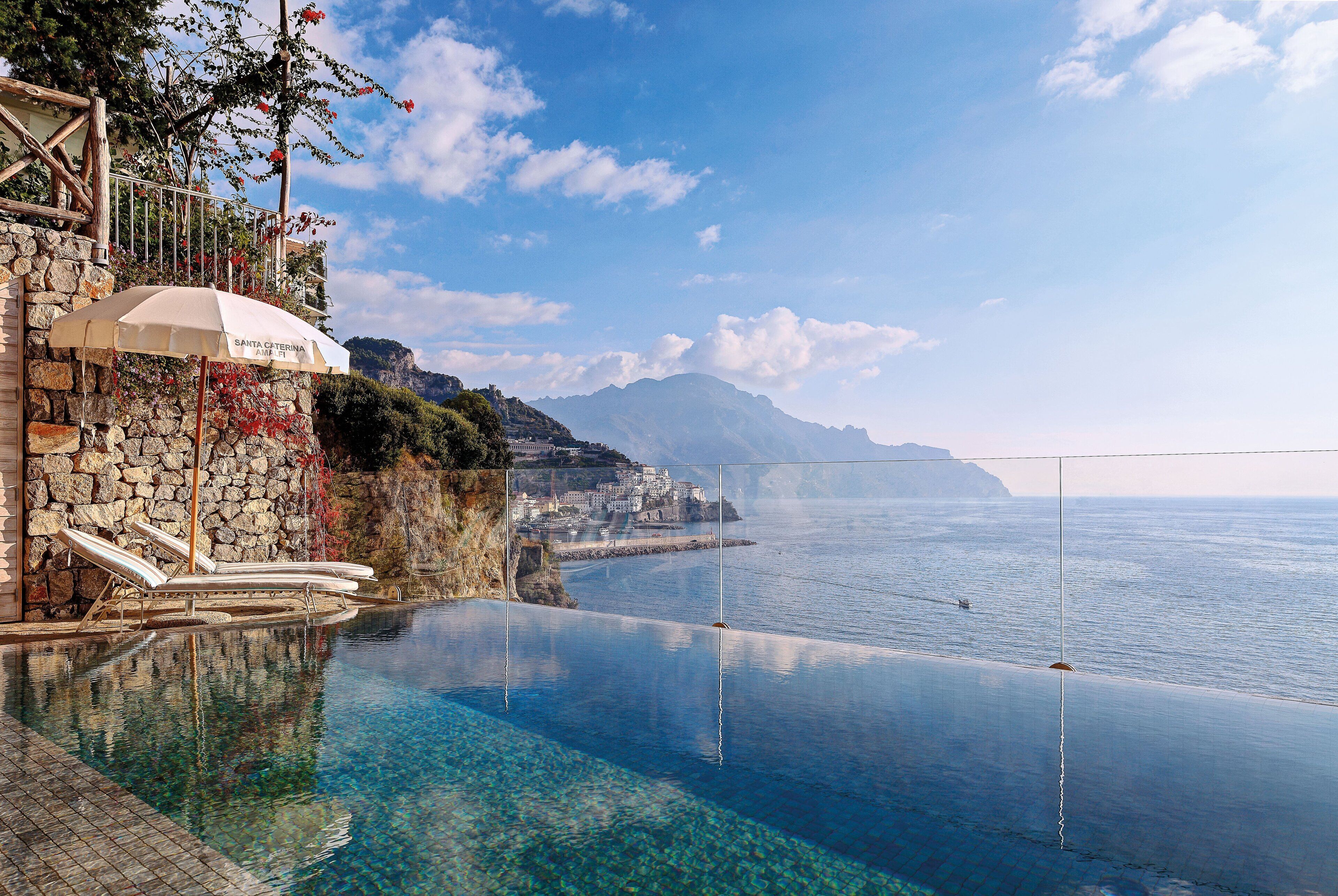 Una piscina infinita con agua cristalina, un muro de piedra y vistas al mar y a las montañas.