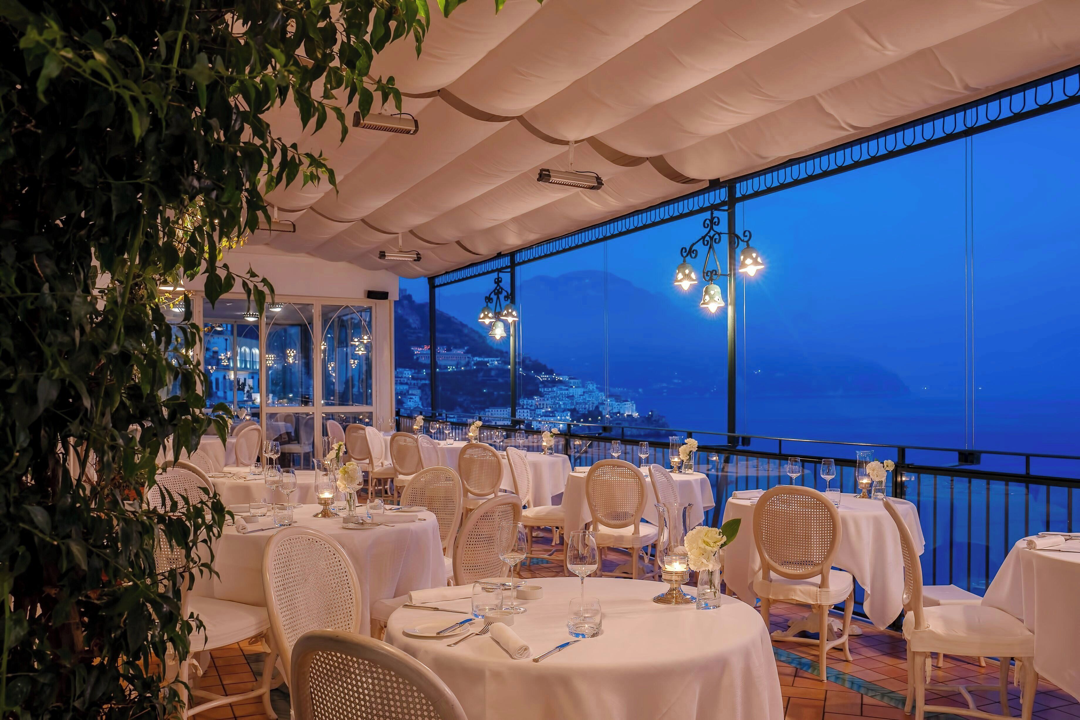 Un restaurant avec des nappes blanches, des chaises en osier et une vue sur la ville au crépuscule.