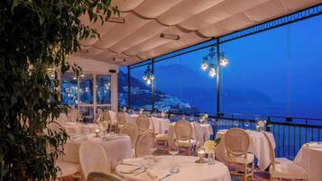 Un restaurant avec des nappes blanches, des chaises en osier et une vue sur la ville au crépuscule.