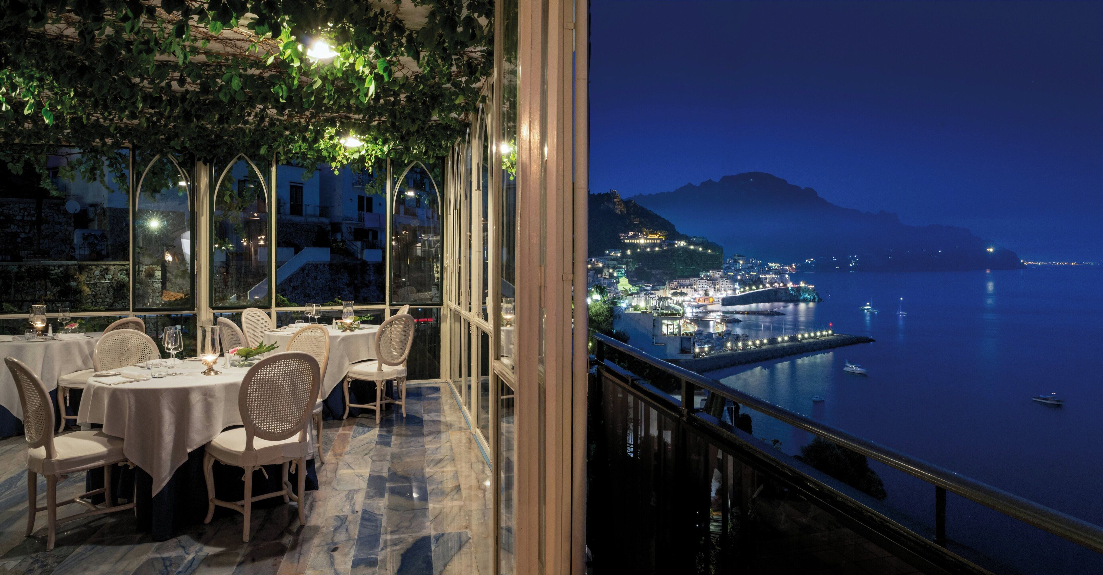 Un restaurant avec des tables et des chaises, une vue sur une ville côtière la nuit, et un plan d’eau.