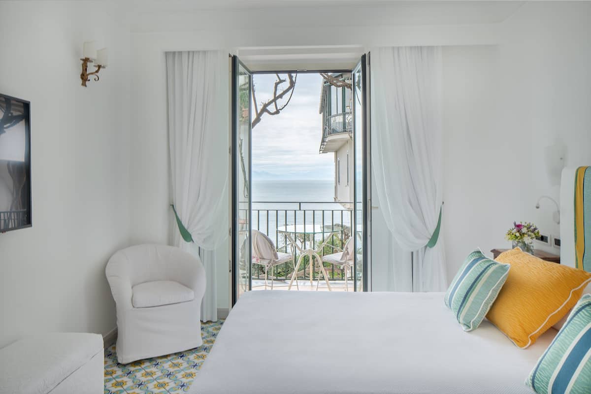 Ein Hotelzimmer mit Bett, Sessel, einem Balkon mit Meerblick und einem Vorhangfenster.
