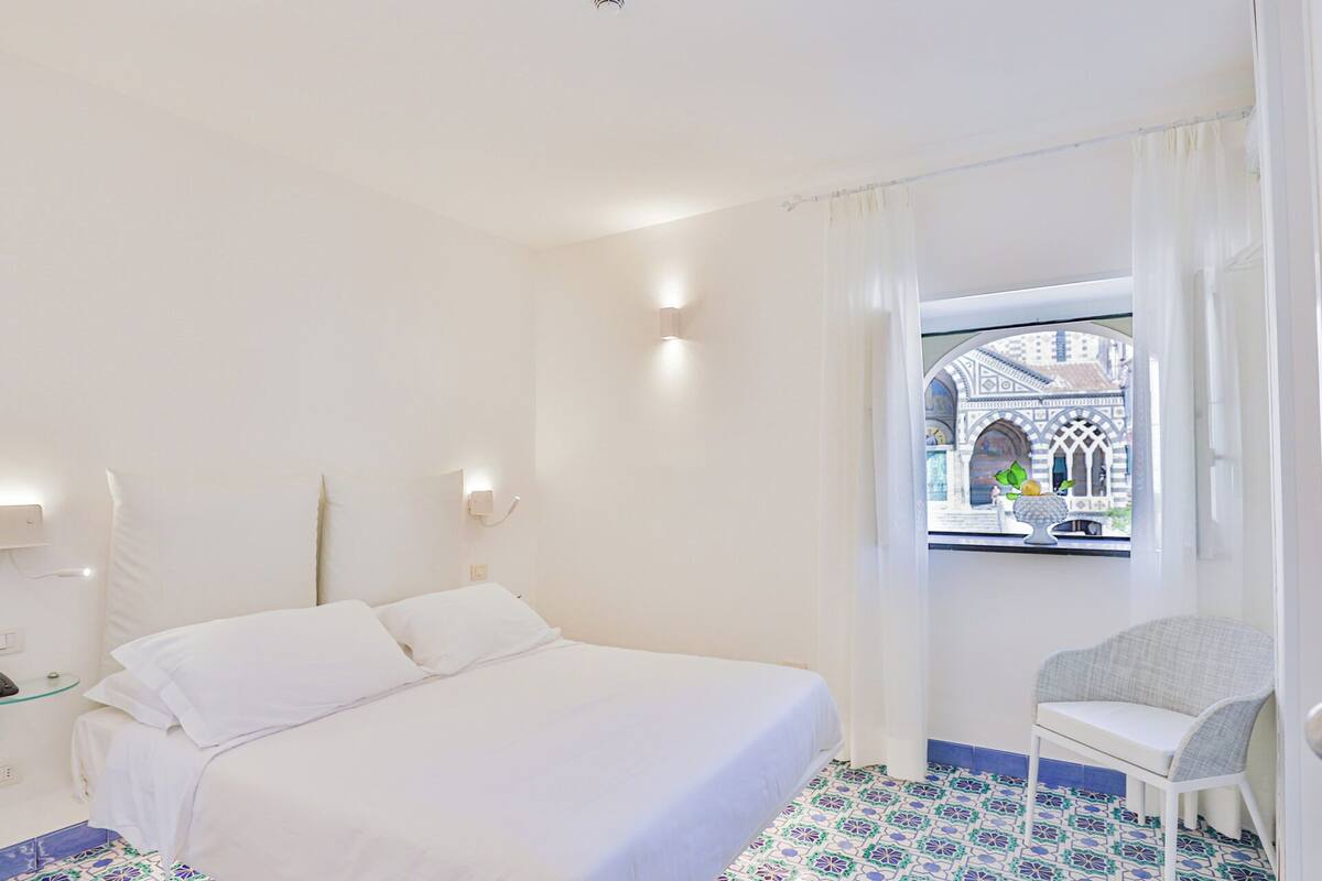 A hotel room with a bed, a chair, a window with a view of a building, and a patterned floor.