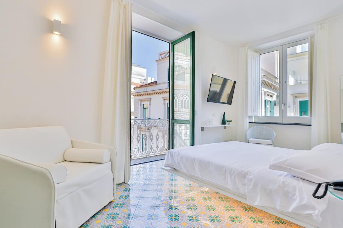 A hotel room with a bed, a chair, a TV mounted on the wall, and a balcony with a view of buildings.
