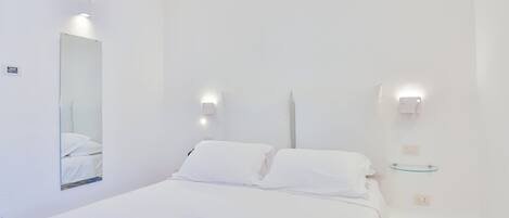 A minimalist hotel room with a large bed, two bedside lamps, a small bedside table, and a mirror.