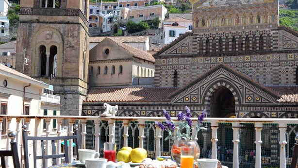 A balcony with a table set for breakfast, overlooking historic buildings and a mountainous backdrop. - Terrazza Duomo (Amalfi)