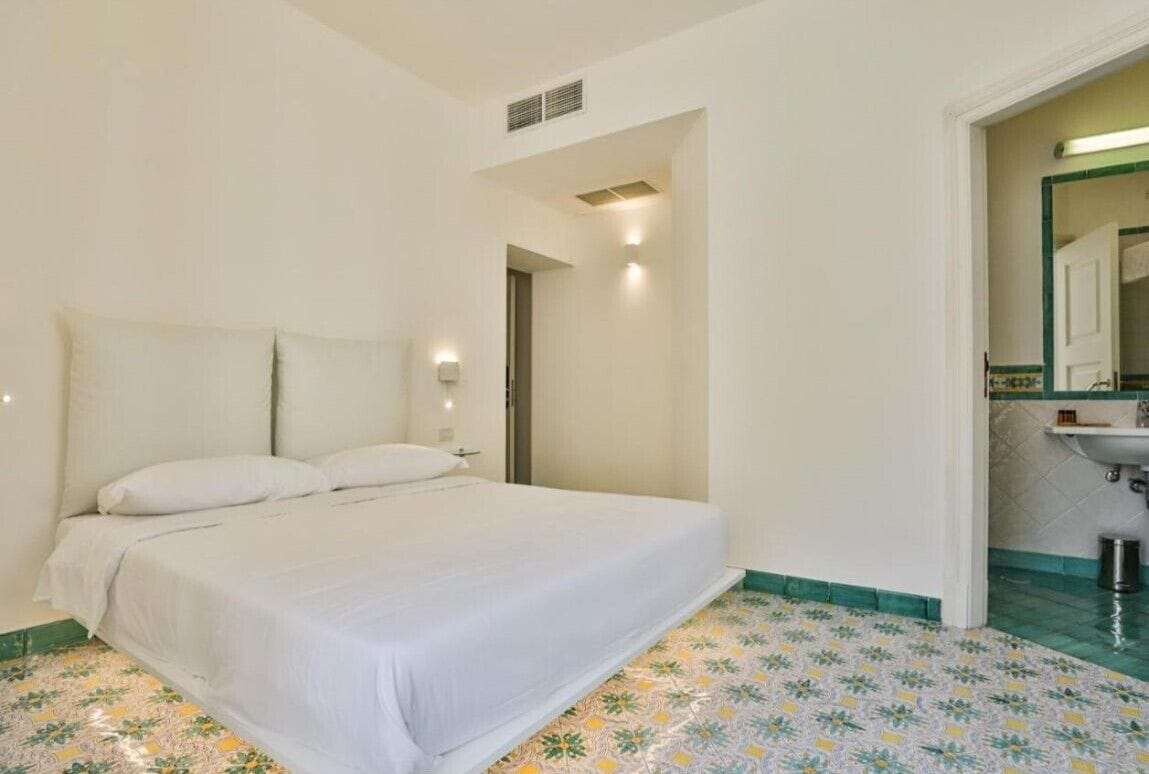 A bedroom with a bed, a nightstand, a wall-mounted light, and a bathroom with a sink and mirror.