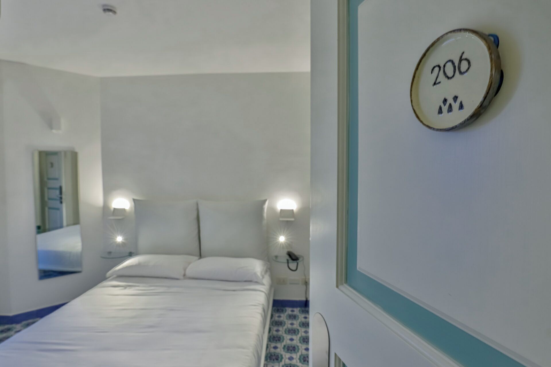 Habitación de hotel con cama, dos lámparas de noche, una placa de número en la pared y suelo estampado.