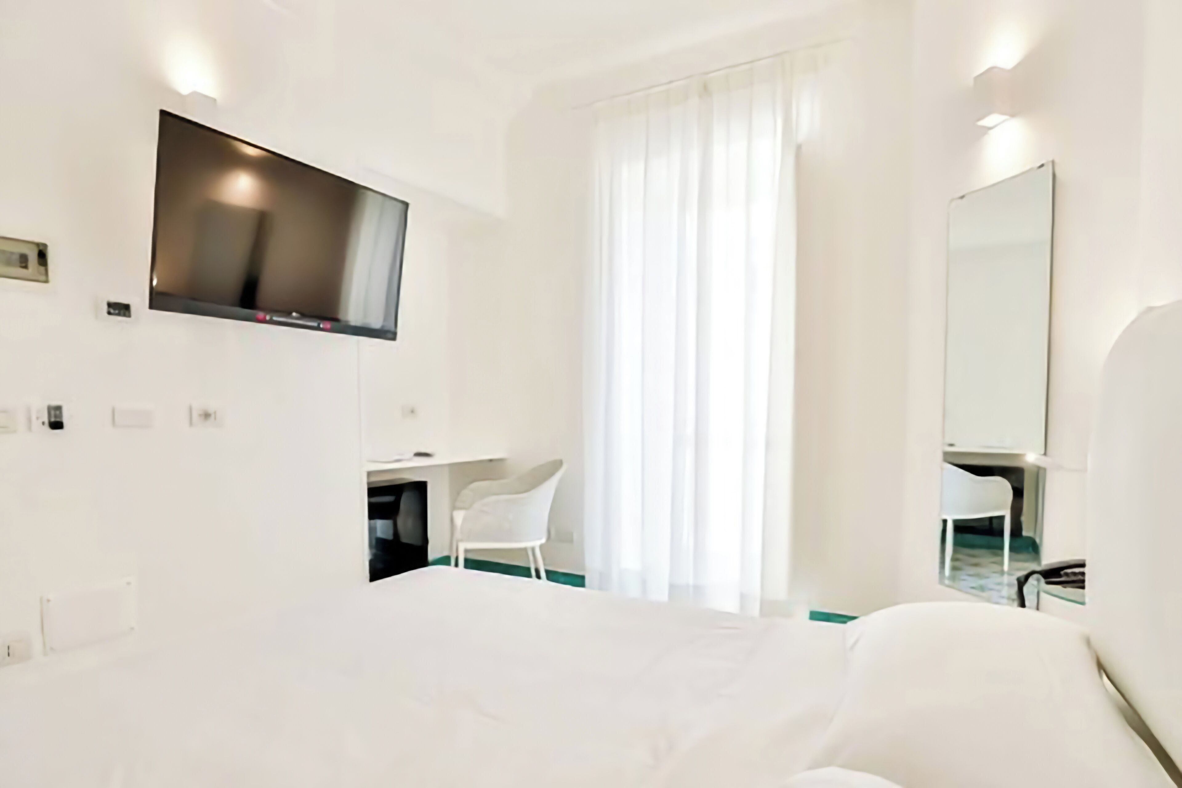 A hotel room with a bed, a desk, a chair, a TV mounted on the wall, and a large window with curtains.