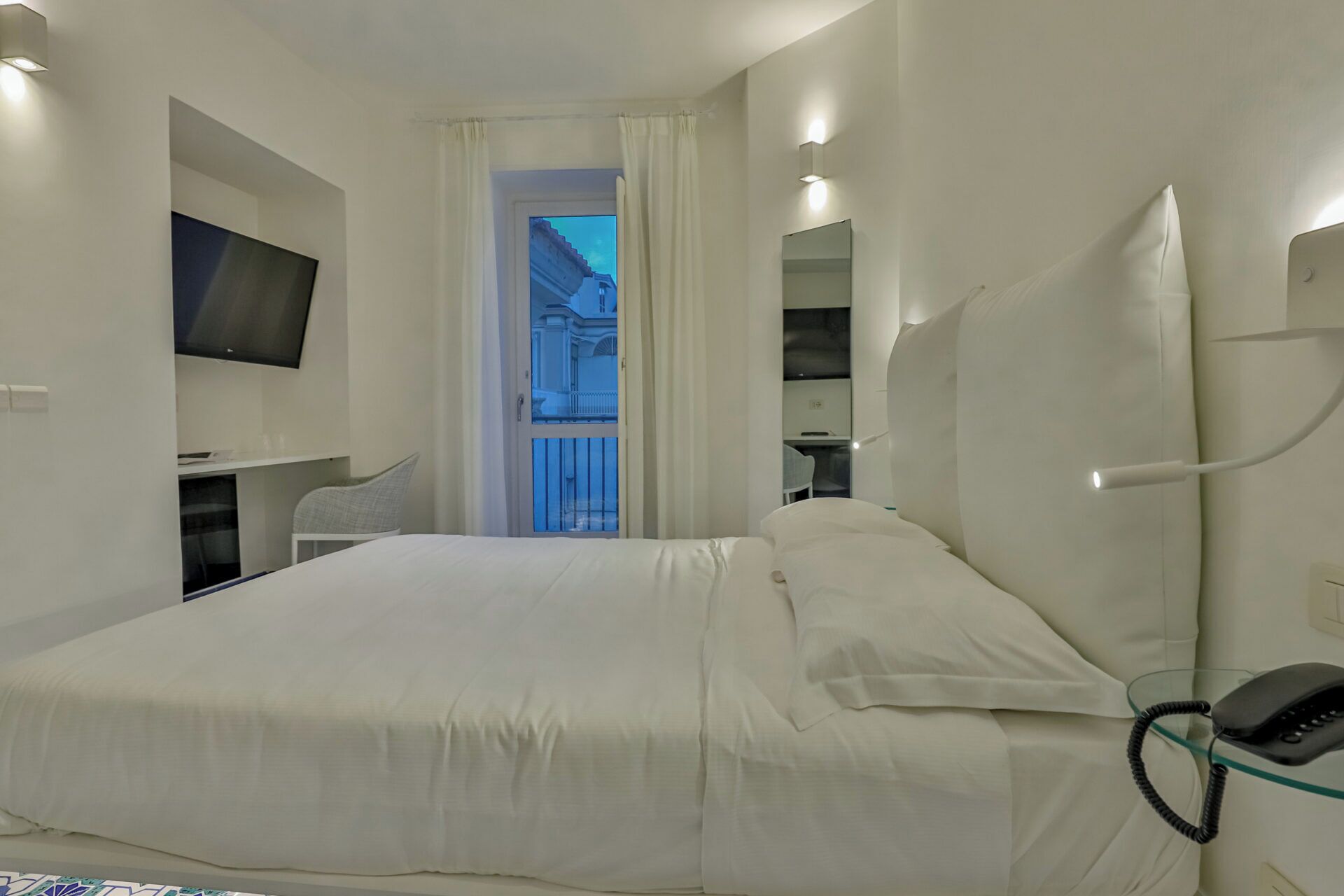 A modern hotel room with a large bed, a TV mounted on the wall, a desk with a chair, and a glass telephone.
