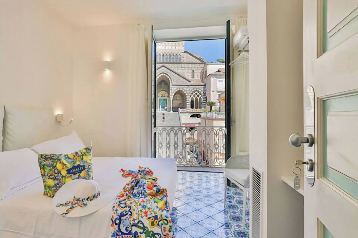 A hotel room with a bed, a colorful pillow, a hat, and a balcony with a view of a historic building.