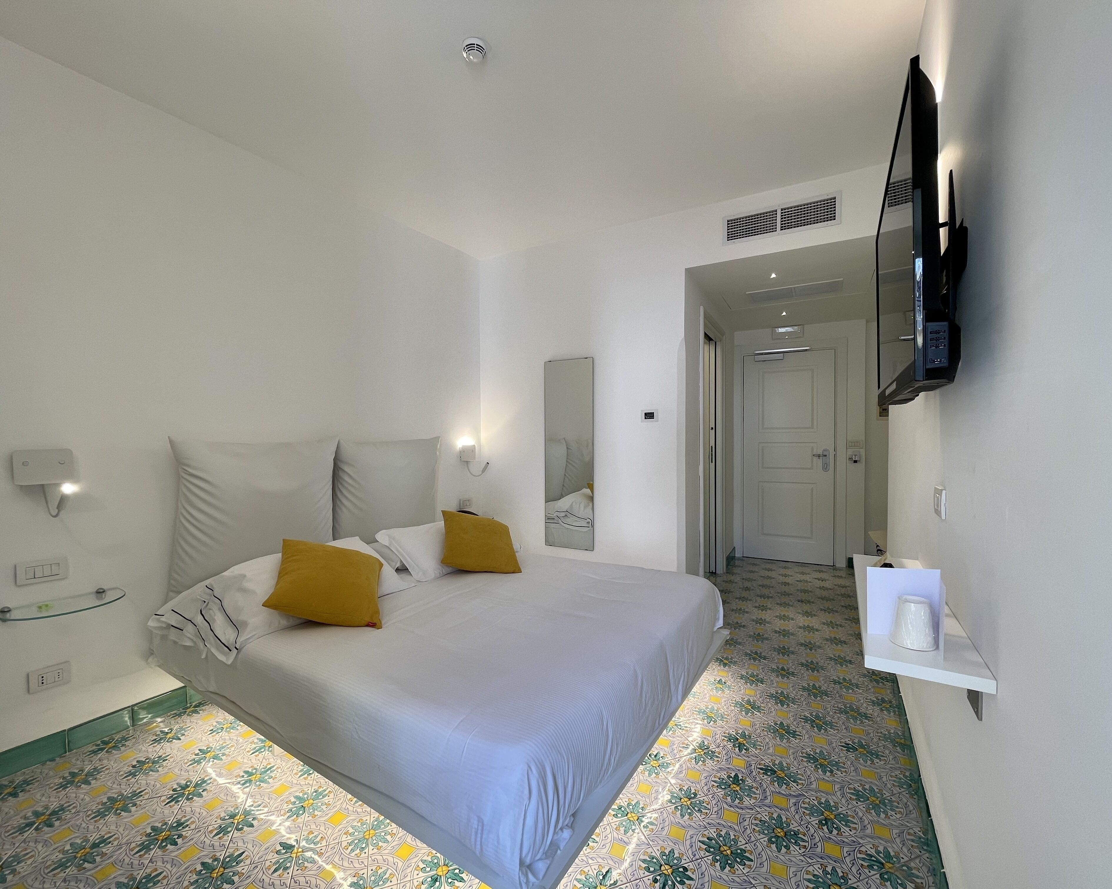 A hotel room with a bed, a TV mounted on the wall, a mirror, and a small table with a cup.