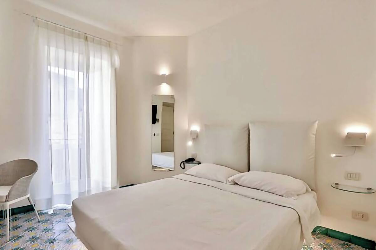 A hotel room with a bed, a chair, a bedside table, a mirror, and a window with curtains.