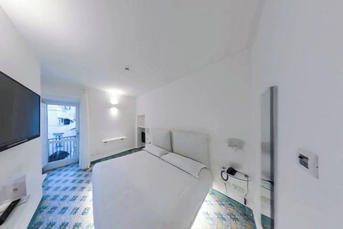 A hotel room with a bed, a flat-screen TV mounted on the wall, a bedside table, and a small window.
